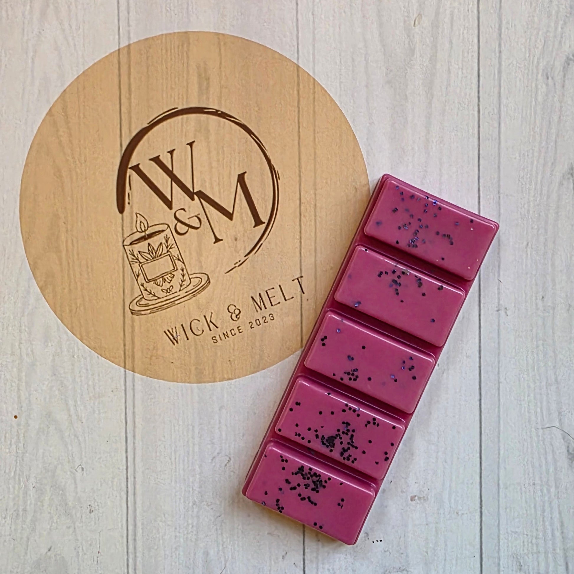 Pink wax bar with glitter on a wooden coaster with a brand logo, on a light wooden surface.