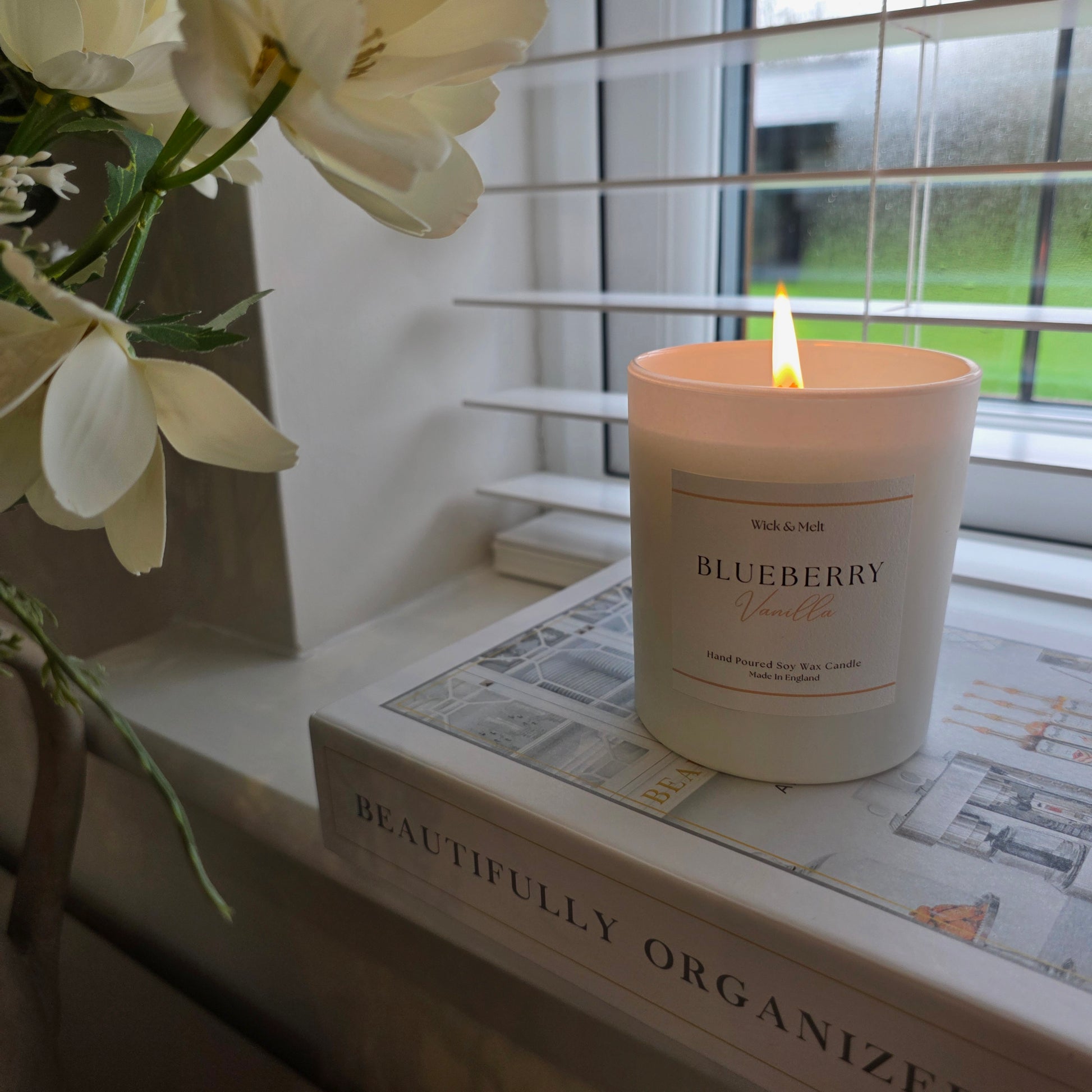 Candle labeled 'Blueberry' on a surface with flowers and a book in the background